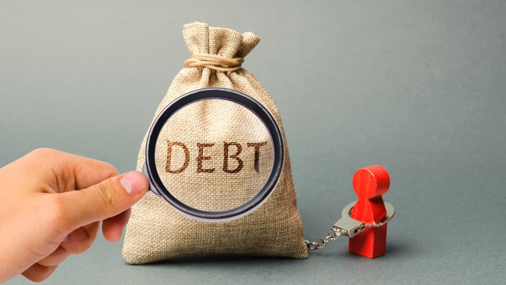 A magnifying glass focuses on a money bag labeled 'Debt,' with a borrower bound by handcuffs. Concept of financial debt scrutiny and entrapment.