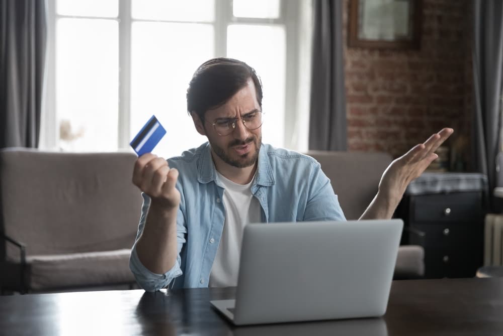 Frustrated man with laptop, reading a message about credit card issues, financial stress, overspending, and potential bankruptcy.
